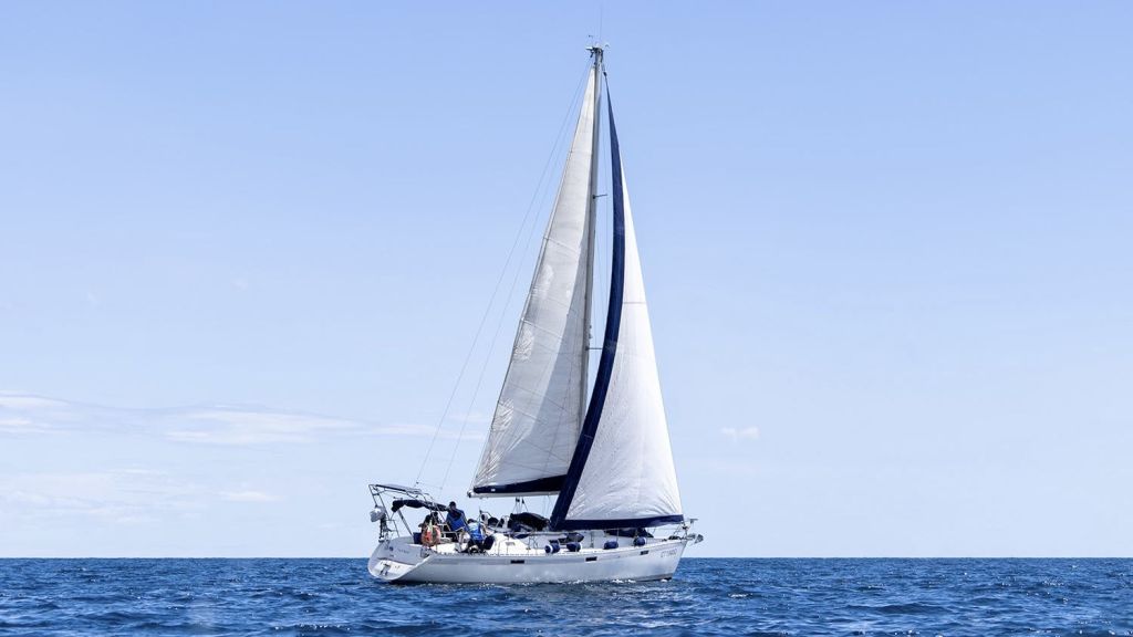 흰색 돛이 달린 범선이 맑고 푸른 하늘 아래 잔잔한 바다를 항해하고 있습니다. 보트 갑판에 사람들이 보입니다.