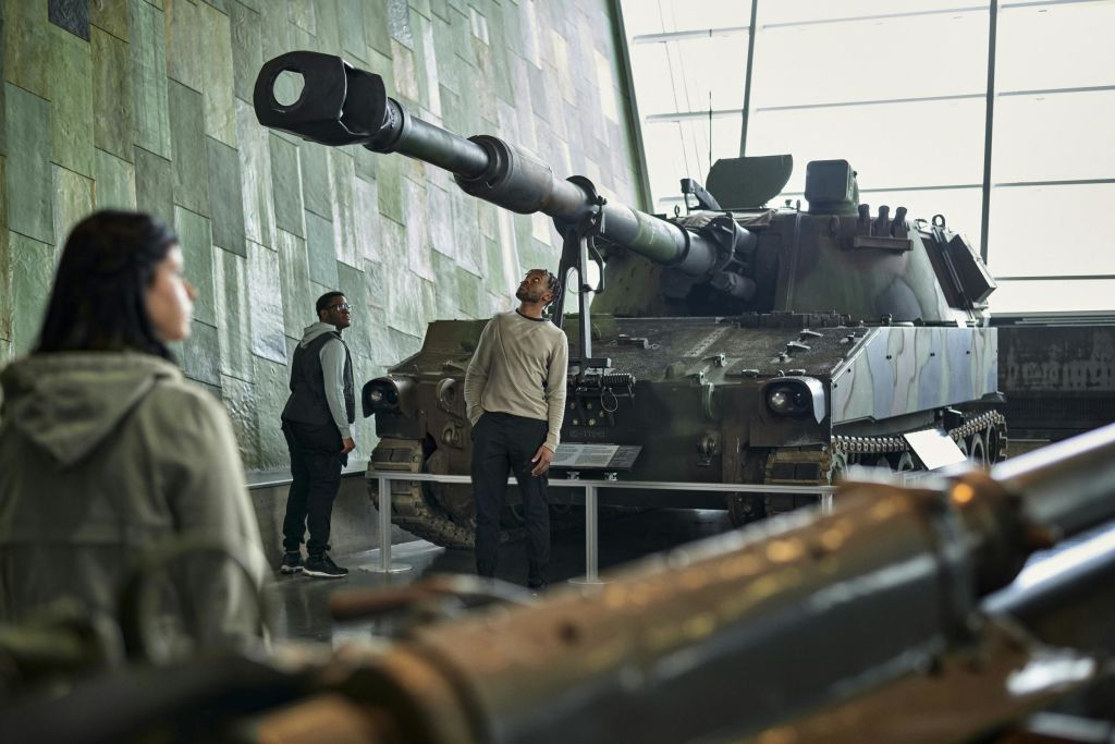 타일로 마감된 벽과 높은 플랫폼이 있는 박물관에서 세 사람이 대형 군사 포병을 살펴보고 있습니다.