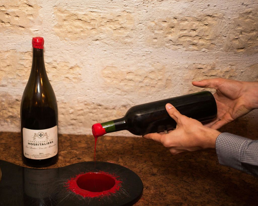 한 사람이 다른 와인병을 들고 있는 테이블 근처에서 레드 왁스로 와인병 목을 돌담에 코팅하고 있습니다.