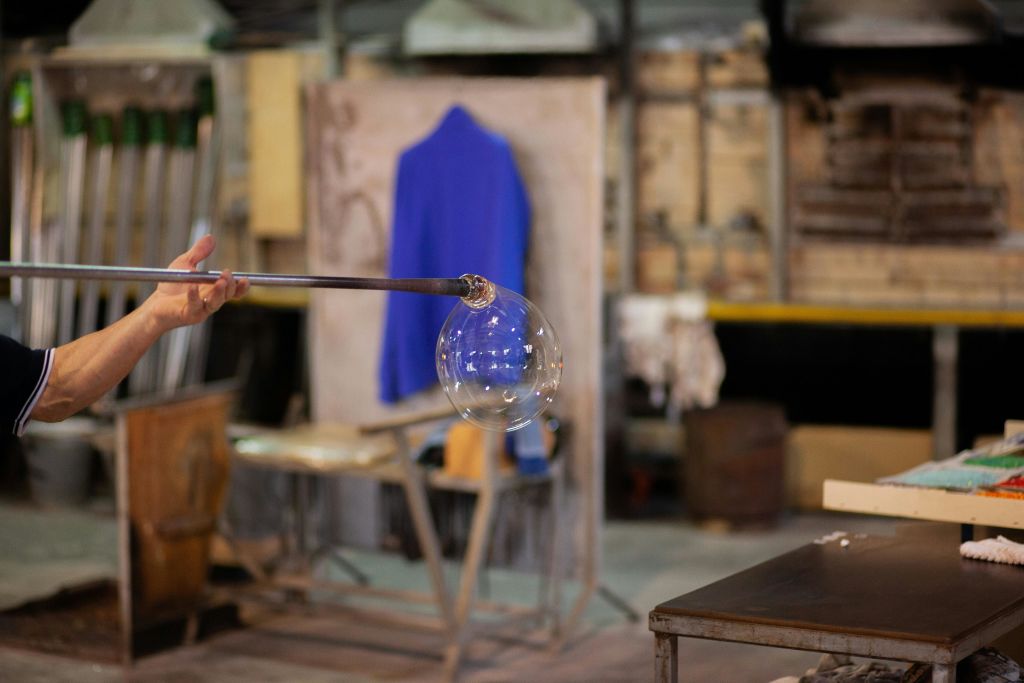 파란색 재킷을 배경으로 워크숍 끝에 유리 거품이 형성된 유리 블로잉 로드를 들고 있는 사람.