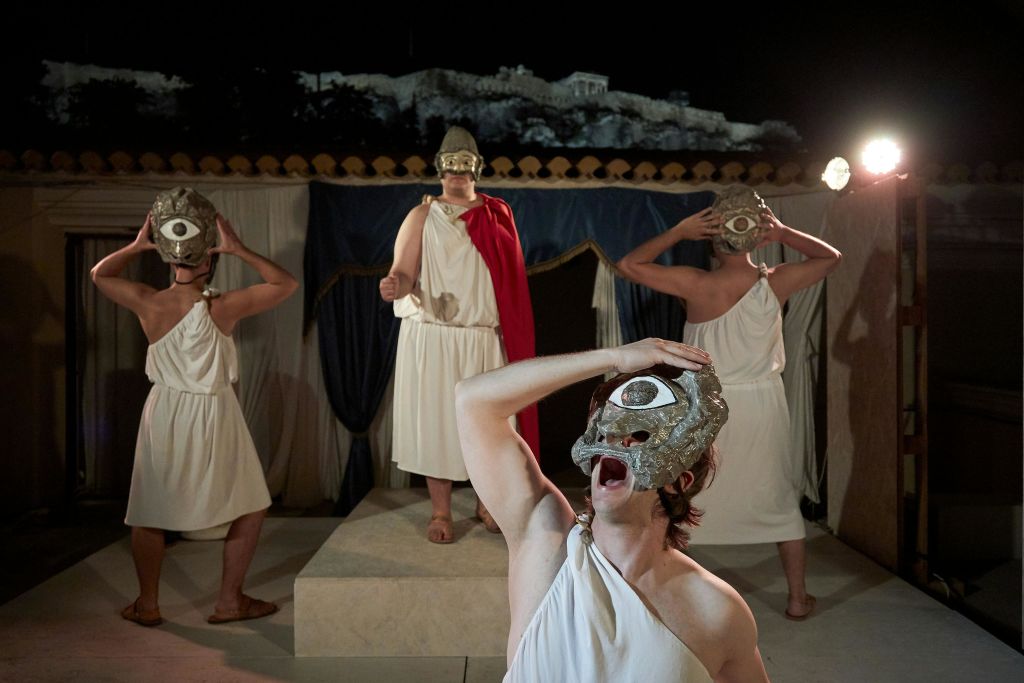 흰색 토가와 아이 마스크를 쓴 네 사람이 커튼과 석조 유적을 배경으로 무대에서 고대 그리스 연극을 공연합니다.