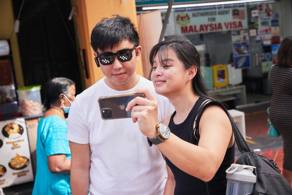 남성과 여성이 셀카를 찍고, 남성은 선글라스를 착용하고, 파란색 드레스를 입은 여성이 배경에 서 있습니다.