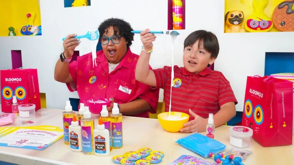 접착제, 그릇, 다채로운 소모품이 놓인 테이블에서 점액을 만드는 두 사람. 하나는 신축성 있는 슬라임을 들고 흥분한 표정입니다.