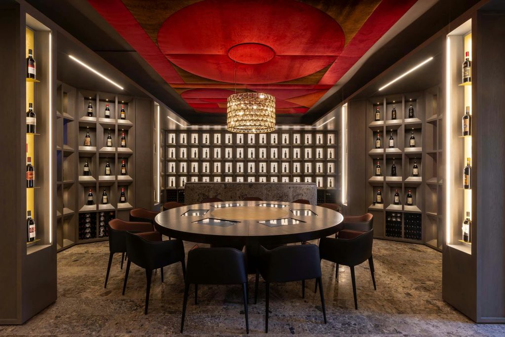 의자, 와인병 선반, 빨간색 천장에 매달린 샹들리에로 둘러싸인 원형 테이블이 있는 현대적인 와인 저장고입니다.