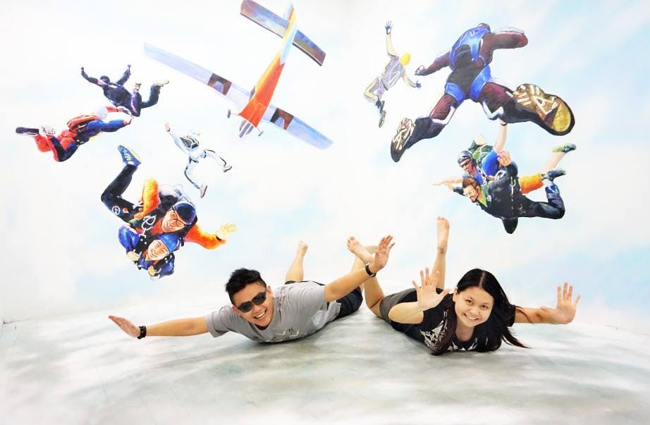 두 사람이 스카이다이빙을 하듯 바닥에서 포즈를 취하고 있는데, 스카이다이버들의 벽화와 벽에 걸린 비행기가 그 뒤에 놓여 있습니다.