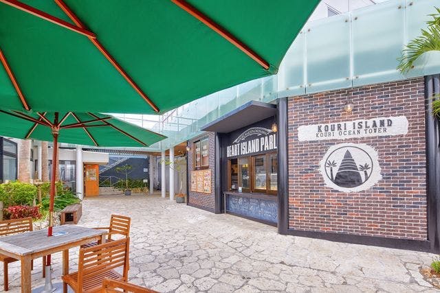초록색 우산, 나무 의자, '하트 아일랜드 팔 (Heart Island Parl)' 이라고 적힌 벽돌로 된 매장이 있는 야외 카페 공간. 간판에는 '코우리섬 바다' 라고 쓰여 있습니다.