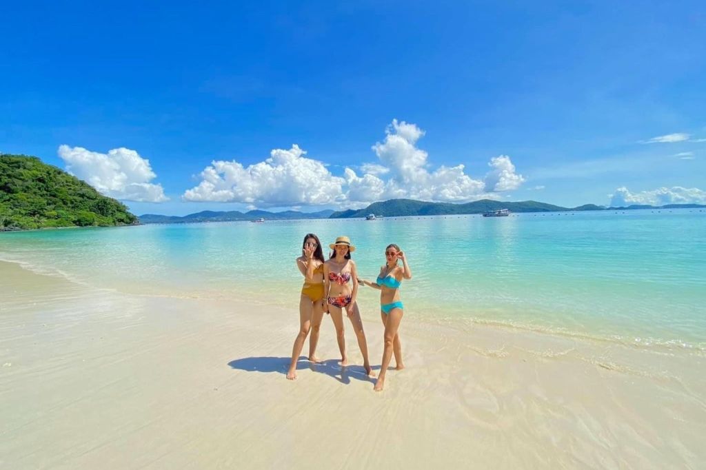 맑은 물과 푹신한 구름이 드리운 푸른 하늘 아래 멀리 떨어진 섬이 있는 햇살 가득한 해변에 수영복을 입은 세 명의 여성이 서 있습니다.