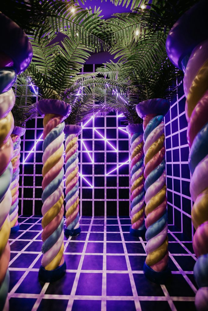 야자수 잎, 네온 보라색 기하학적 조명, 체크무늬 흑백 바닥과 벽이 있는 다채로운 나선형 기둥.