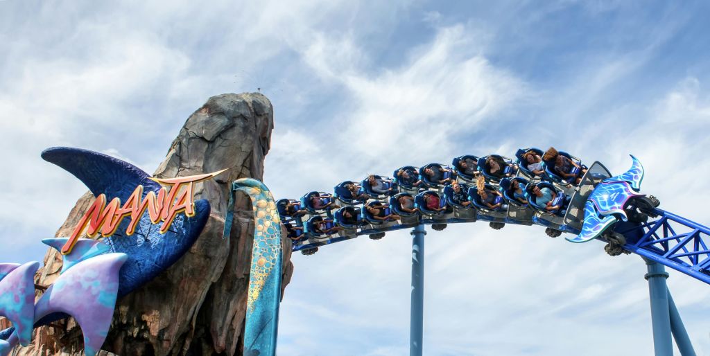 부분적으로 흐린 하늘 아래, 푸른색 롤러코스터를 타고 주제별 장식으로 장식된 바위 구조물을 둘러싸고 있는 사람들.