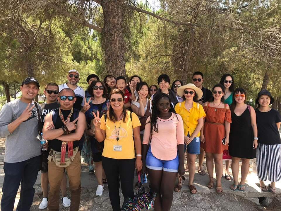 나무 아래에서 포즈를 취하는 다양한 무리의 사람들, 햇살이 내리쬐는 야외 환경에서 많은 사람들이 미소 짓고 평화 표지판을 만들고 있습니다.