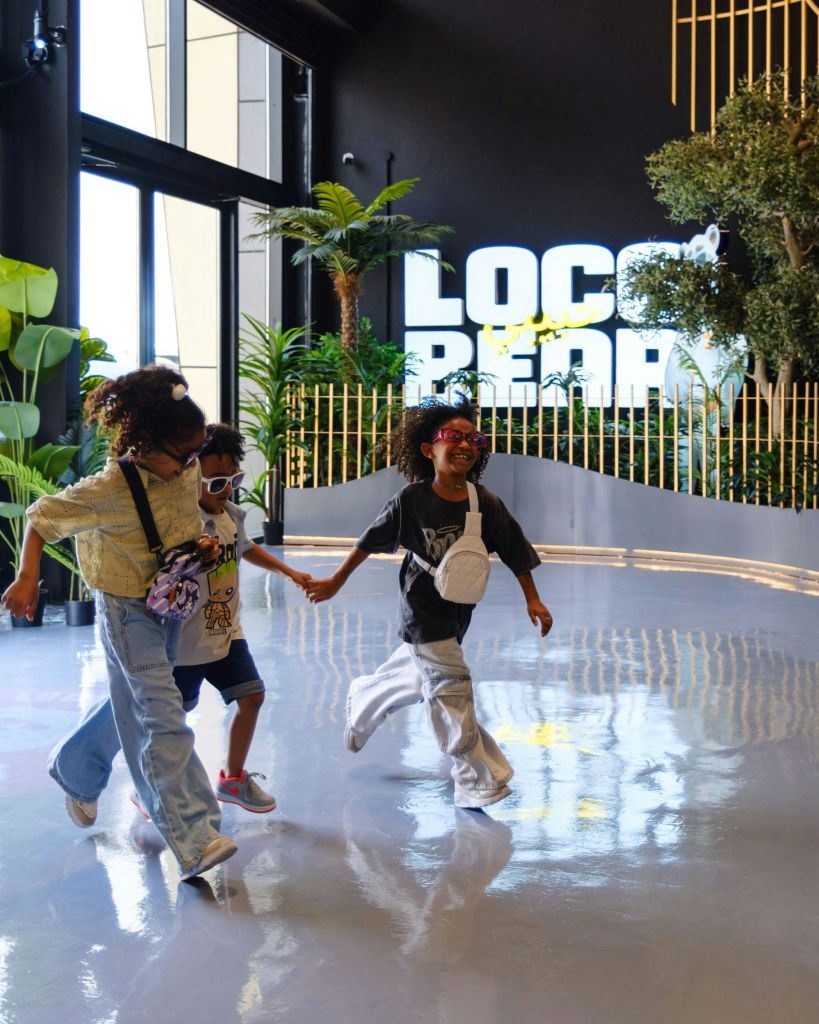 캐주얼한 옷과 선글라스를 입은 세 명의 어린이가 "Loco Pez" 간판이 있는 현대적이고 열대 테마의 공간에서 함께 달립니다.