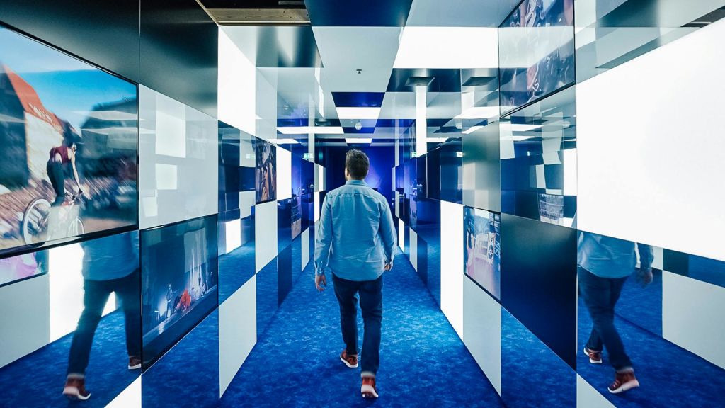 파란색 셔츠를 입은 사람이 스크린과 거울이 있는 체크무늬 벽이 있는 파란색 카펫이 깔린 복도를 걸어가고 있습니다.