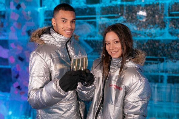 은색 파카를 입은 미소 짓는 커플이 얼음 벽 앞에 서서 샴페인 잔을 들고 있습니다.