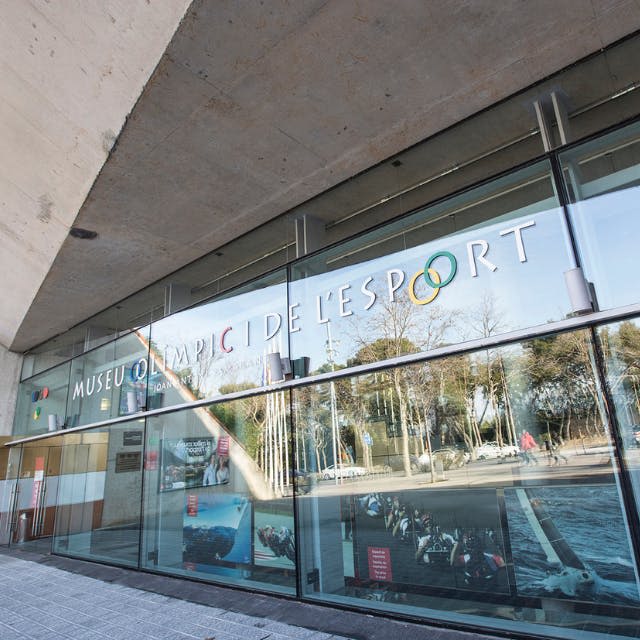 "Museu Olímpic de l 'Esport" 표지판이 있는 커다란 유리 창문이 있는 현대적인 건물로 밖에 나무가 반사되어 있습니다.