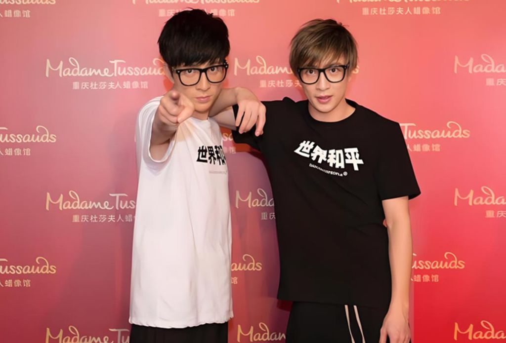 안경을 쓰고 테마가 있는 티셔츠를 입은 두 사람이 마담 투소 로고가 있는 빨간색 배경 앞에서 포즈를 취하고 있습니다.