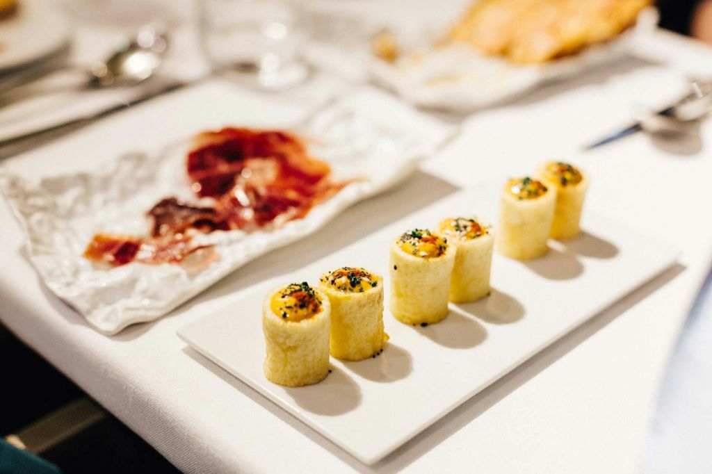 흰색 직사각형 접시에 네 개의 스시 롤이 깔끔하게 배열되어 있으며, 얇게 썬 고기 접시가 배경에 있습니다.