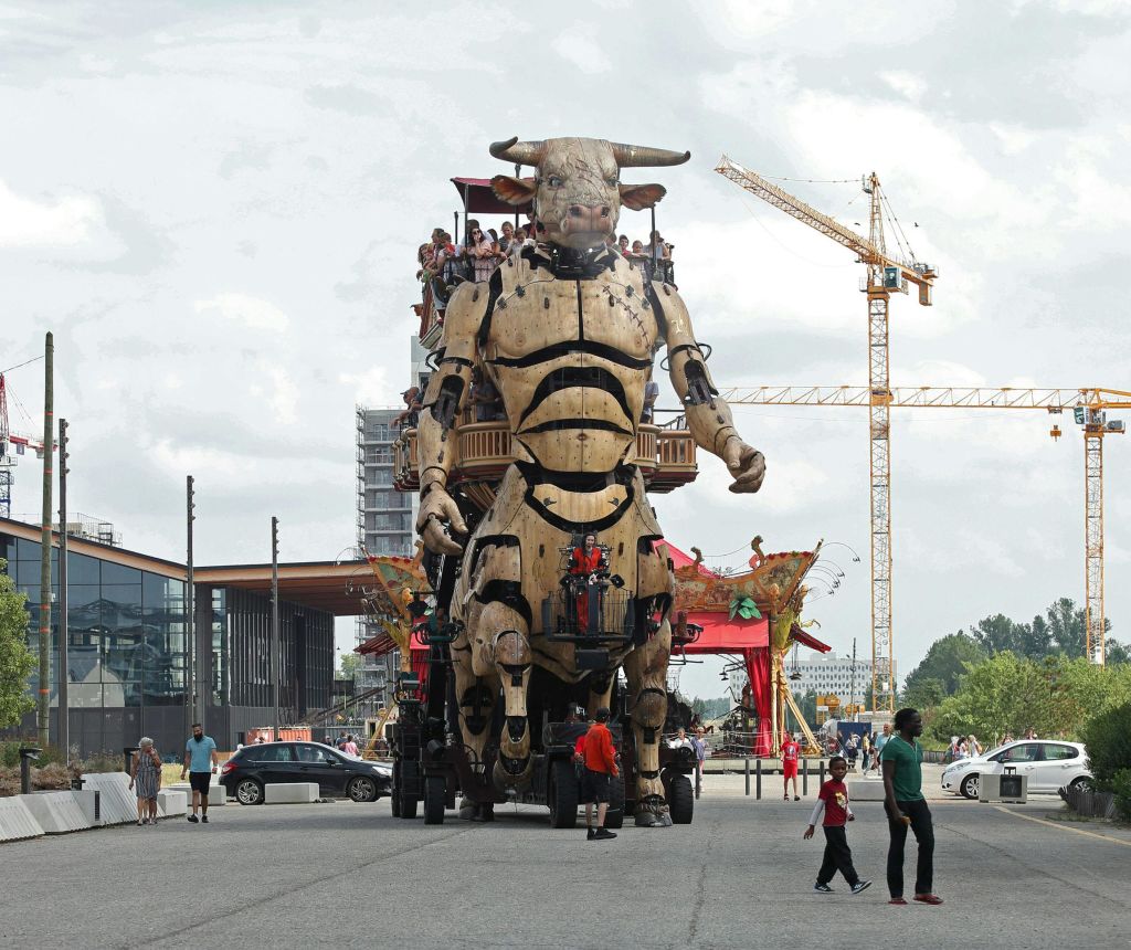 사람들을 등에 업은 커다란 기계식 황소가 거리를 지나갑니다. 구경꾼, 크레인, 건물, 자동차를 배경으로 합니다.