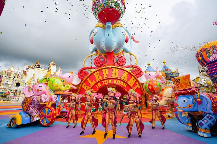 화려한 의상을 입은 연주자들이 활기찬 "Elephant in the Room" 카니발 전시장 앞에서 춤을 추고 있습니다. 콘페티는 위에서 떨어집니다.