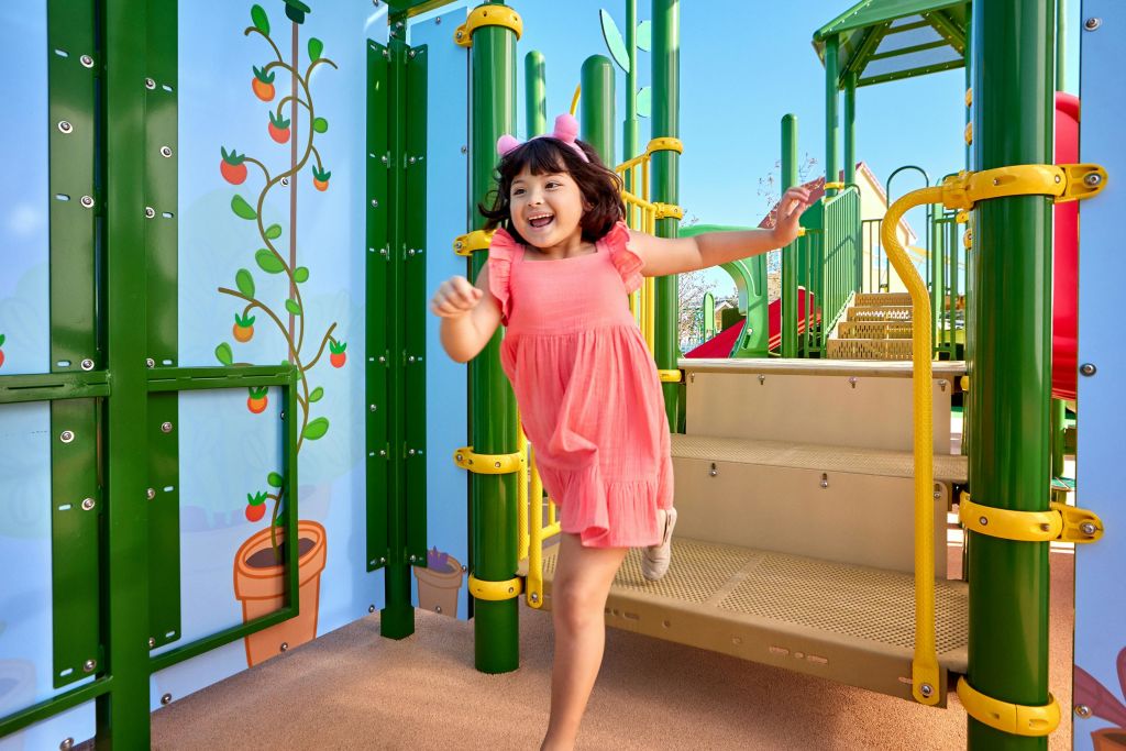 분홍색 드레스와 머리띠를 한 어린 소녀가 녹색 구조물과 계단이 있는 다채로운 운동장에서 즐겁게 달리고 있습니다.