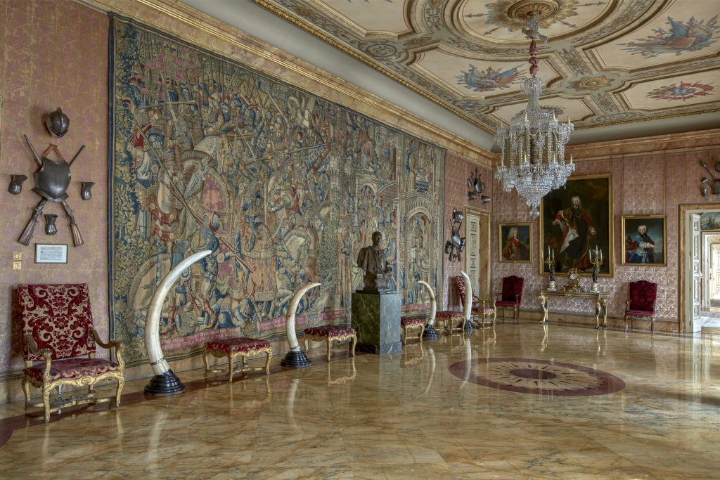 커다란 벽면 태피스트리, 대리석 바닥, 샹들리에, 흉상 조각, 의자와 벤치를 포함한 앤티크 가구가 있는 화려한 객실입니다.