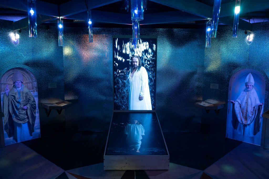 파란색 조명이 있는 반짝이는 금속으로 된 방에 세워진 흰색 가운을 입은 사람이 반사되어 있는 디스플레이.