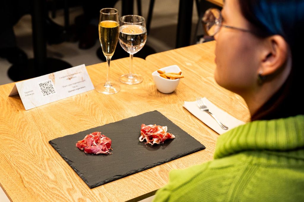 녹색 스웨터를 입은 사람이 경화 고기 한 판, 음료수 한 잔, 브레드스틱 한 그릇이 놓인 테이블에 앉아 있습니다.