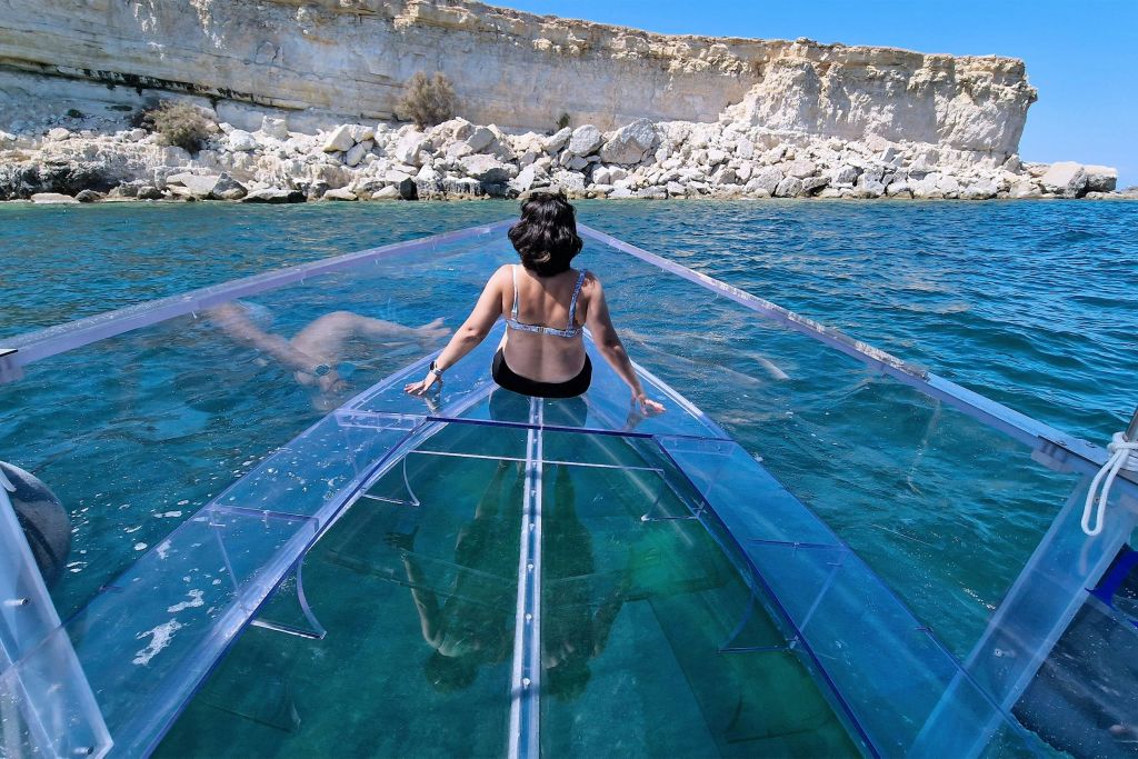 수영복을 입은 한 사람이 햇살이 내리쬐는 하늘 아래 바위 해변 근처의 맑고 푸른 바다에서 투명한 보트에 앉아 있습니다.