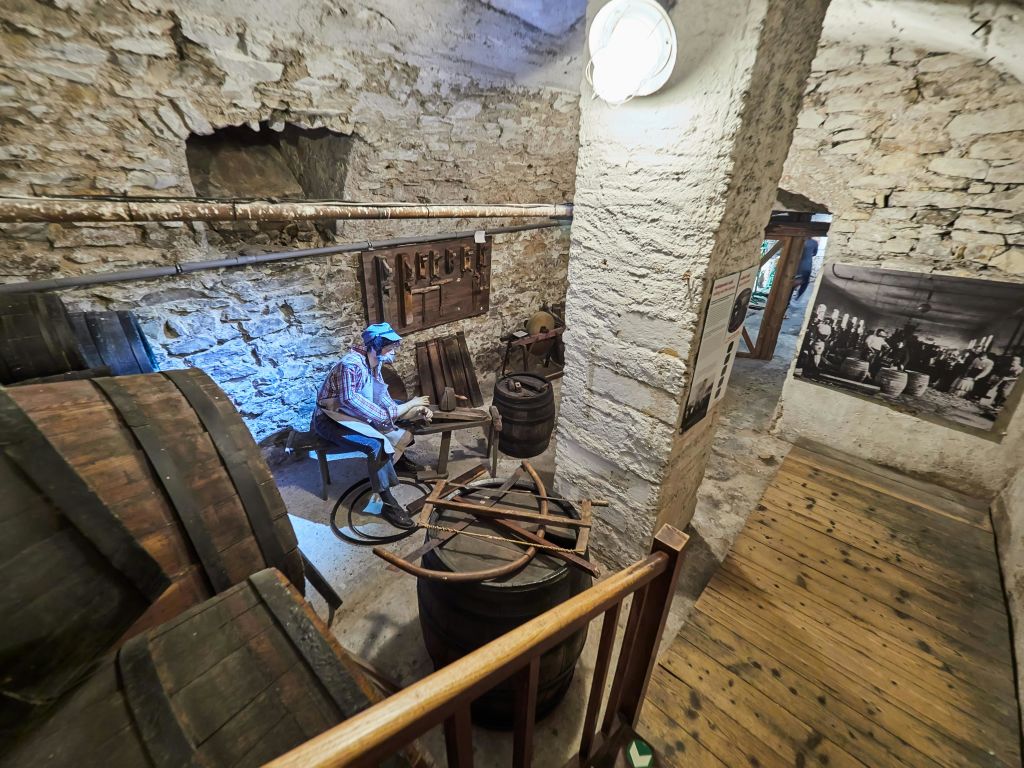 소박한 방의 디오라마에는 나무 통과 도구로 작업하는 마네킹이 있으며, 돌담과 나무 바닥 옆에 있습니다.