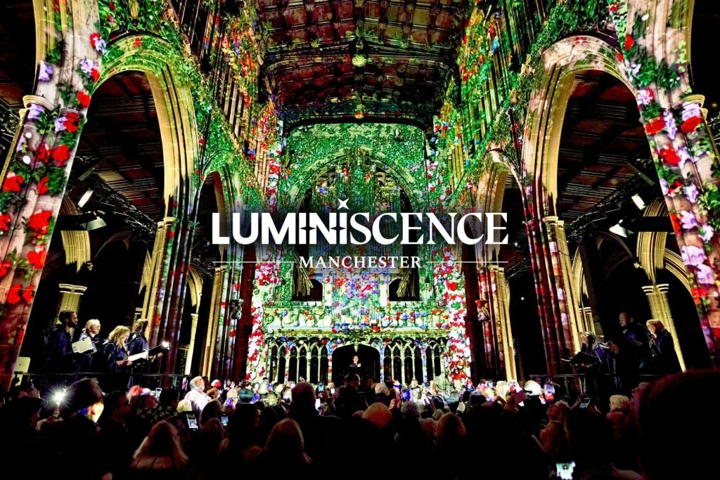역사적인 건물 내부에서 관객과 함께 생동감 넘치는 조명 투영 쇼가 펼쳐집니다. "Luminescence Manchester" 라는 문구가 적혀 있습니다.
