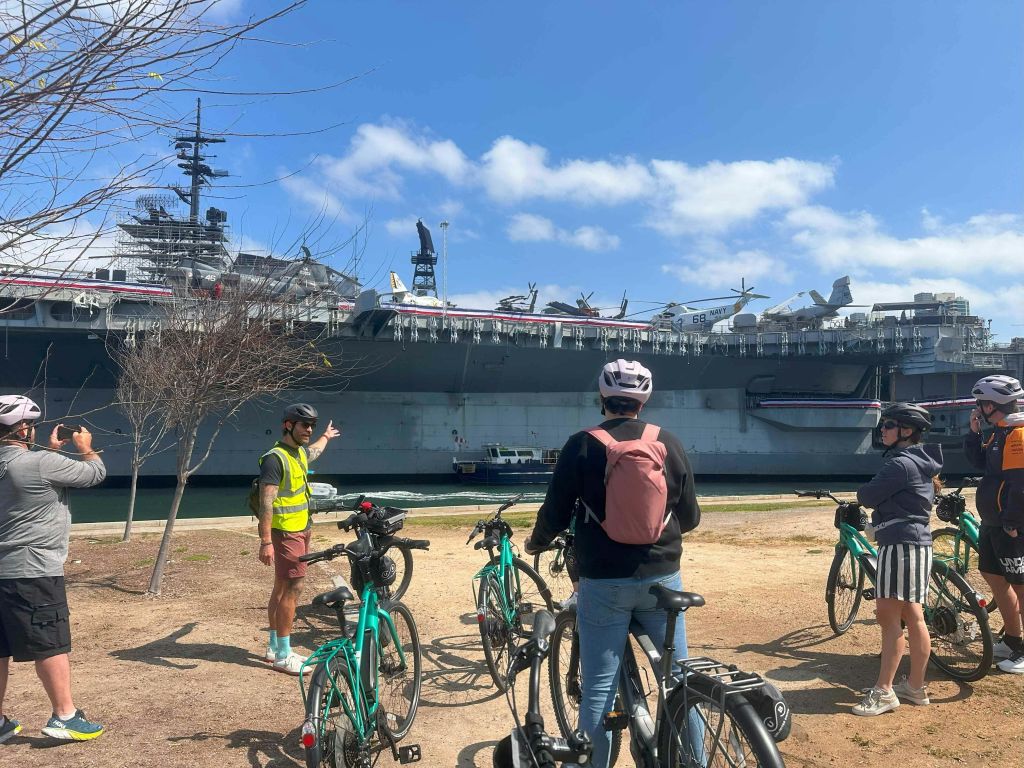 한 무리의 자전거 운전자들이 구름이 흩날리는 맑고 푸른 하늘 아래 도킹된 항공모함을 바라보고 있습니다.