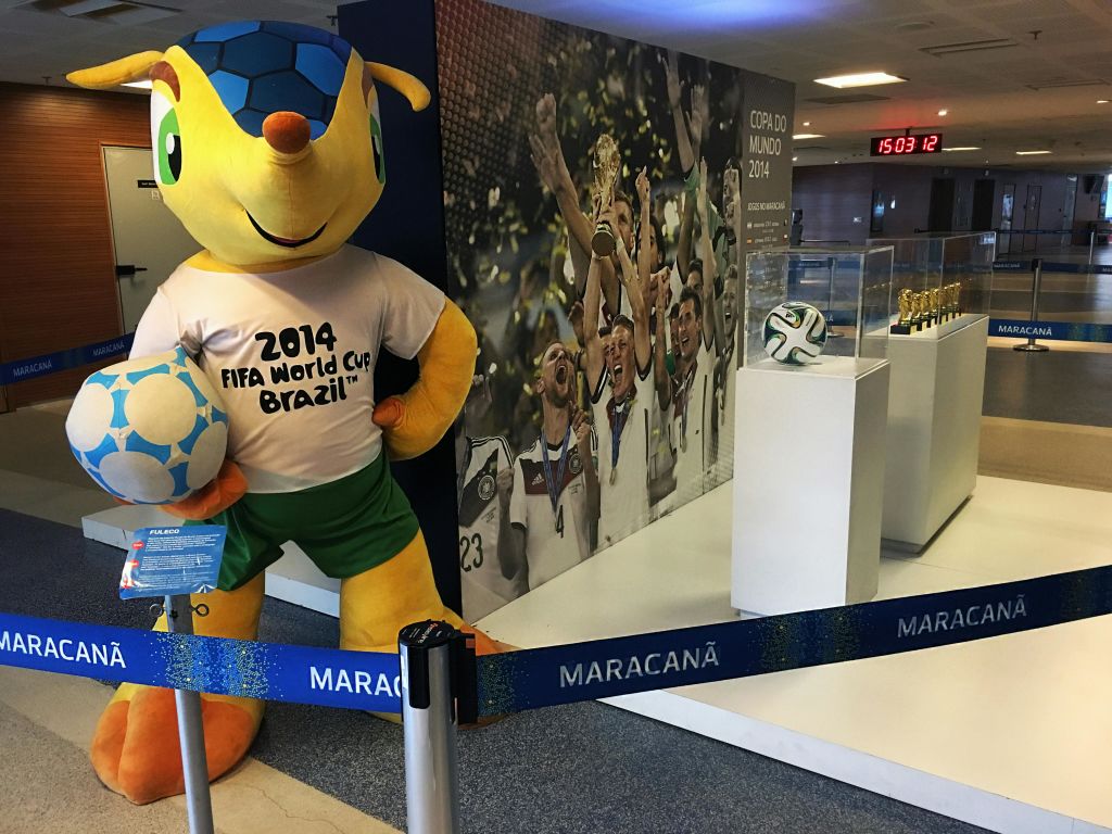 2014 FIFA 월드컵 마스코트가 마라카낭 스타디움에서 우승을 축하하는 선수들의 사진과 트로피 전시 옆에 서 있습니다.