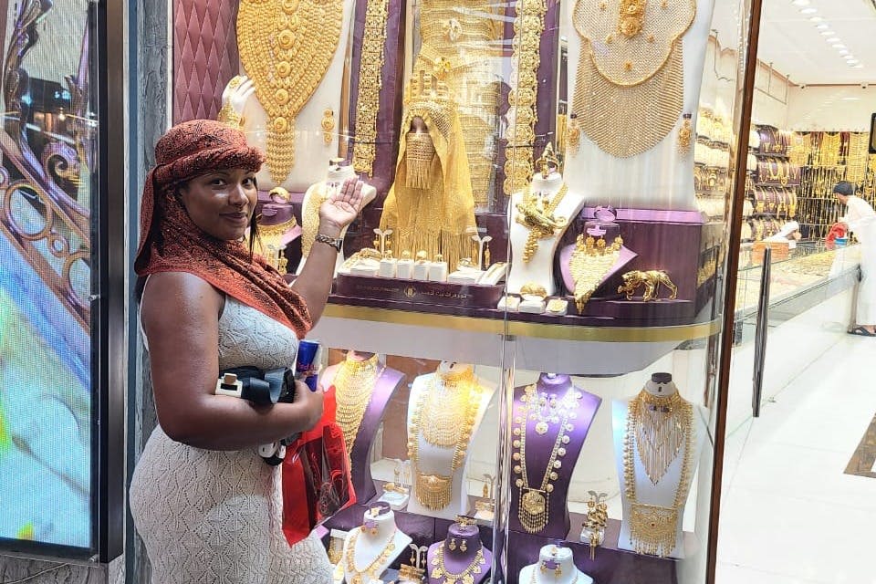 가벼운 드레스를 입고 머리에 스카프를 두른 한 여성이 상점 창문 앞에 서서 다양한 금 보석 조각을 전시하고 있습니다.
