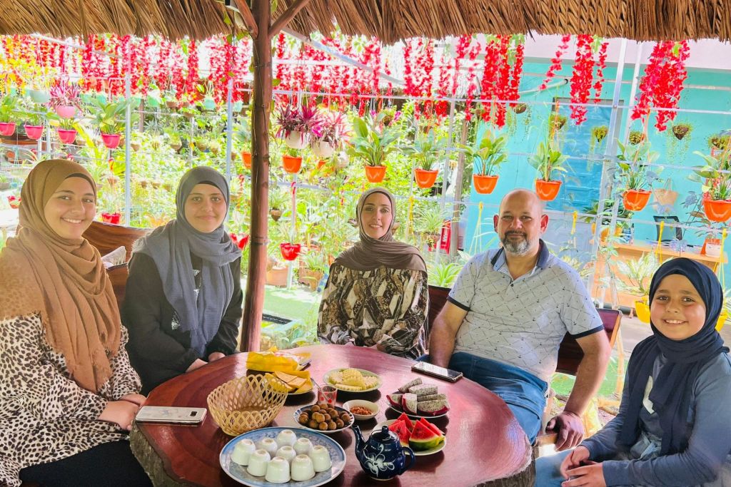세 명의 여성이 둥근 테이블에 앉아 음식과 차를 마시고 초가 지붕 아래 화려한 걸이 식물로 둘러싸여 있습니다.