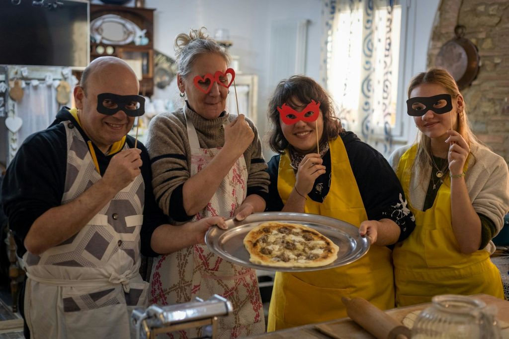 앞치마를 두른 세 사람이 마스크 소품을 들고 있습니다. 한 여성이 트레이에 피자를 선물합니다. 밝은 조명이 있는 주방 설정.