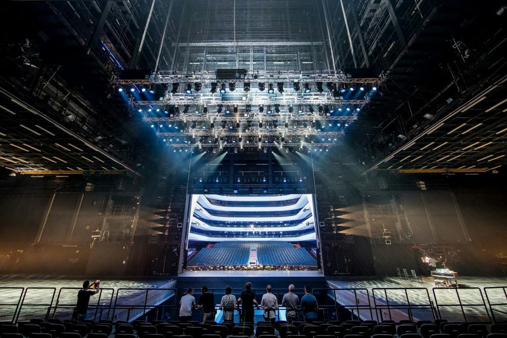 6명으로 구성된 그룹이 대형 극장의 빈 무대 앞에 서서 조명이 켜진 무대와 줄지어 있는 객석 좌석을 마주하고 있습니다.