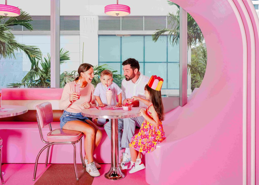 분홍색 부스, 원형 테이블, 창문을 통해 보이는 야자수가 있는 분홍색 테마의 식당에서 4인 가족이 음료를 즐기고 있습니다.