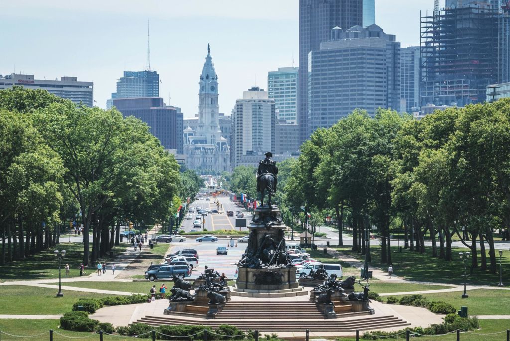 공원 길 끝에 커다란 동상이 서 있으며, 분주한 도시 거리와 고층 빌딩을 배경으로 서 있습니다.