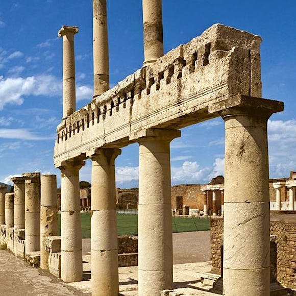 구름이 흩어져 있고 부분적인 구조물이 배경으로 보이는 푸른 하늘 아래 고대 석조 기둥과 유적.