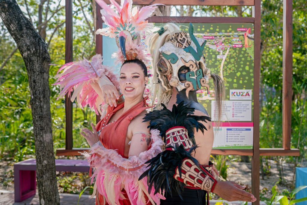 깃털과 마스크가 달린 정교한 의상을 입은 두 사람이 야외에서 나무와 표지판을 배경으로 서 있습니다.