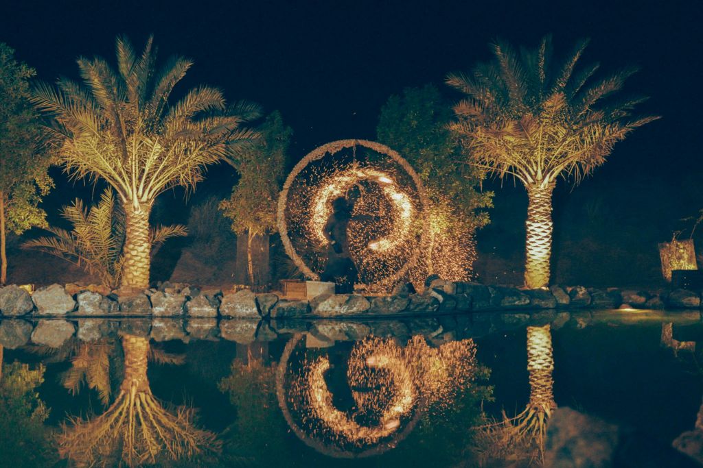 야자수가 우거진 야간 무대에서 공연자가 불을 뿜으며 잔잔한 물결을 반사합니다.