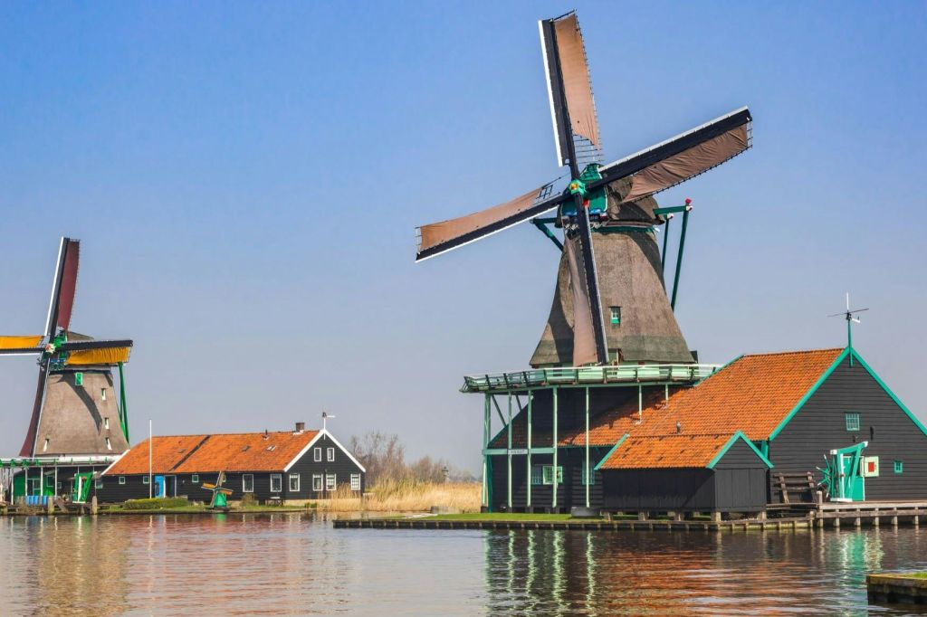 커다란 칼날이 달린 풍차가 맑은 날 잔잔한 수로 옆 붉은 지붕의 건물 옆에 있습니다.