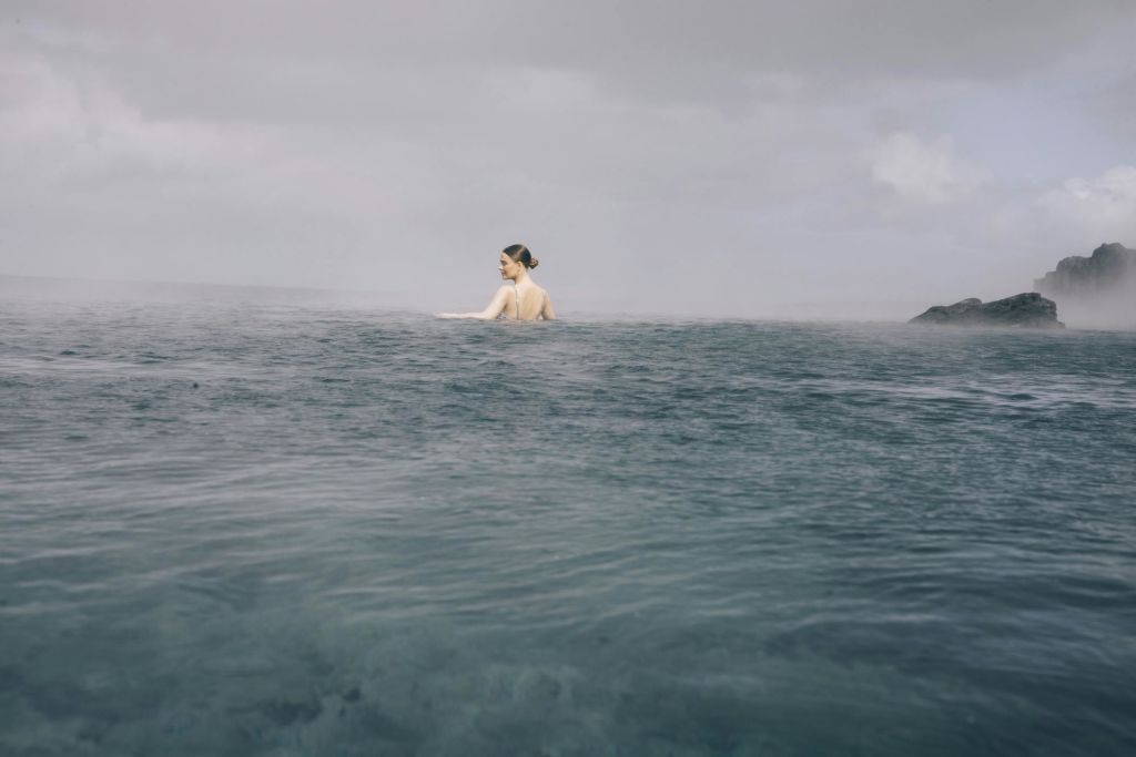 멀리 해안선이 거의 보이지 않는 흐린 하늘 아래 잔잔한 바닷물에서 한 사람이 홀로 수영하고 있습니다.