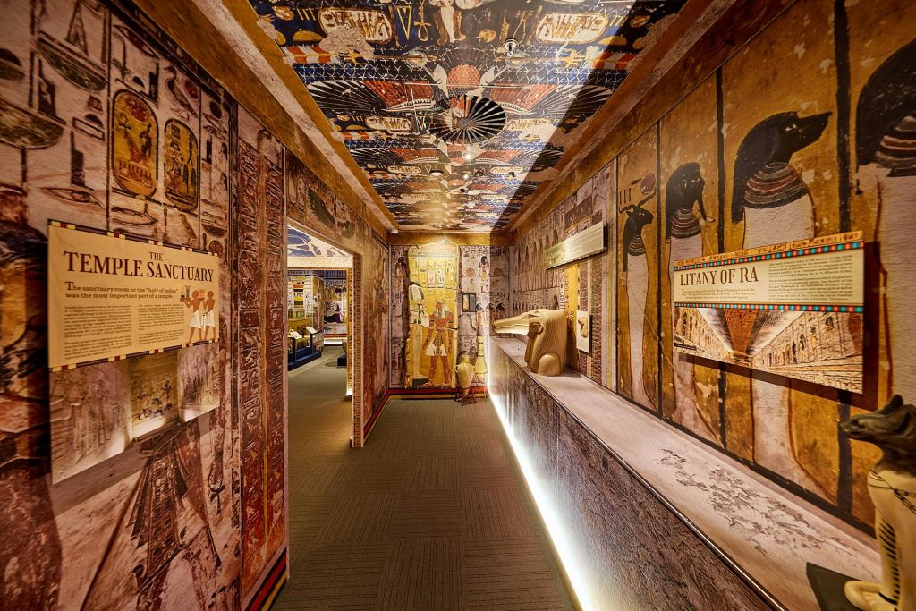 상형 문자로 덮인 벽과 천장이 있는 이집트 테마의 박물관 복도에는 유물, 조명 디스플레이, 대형 동상이 있습니다.