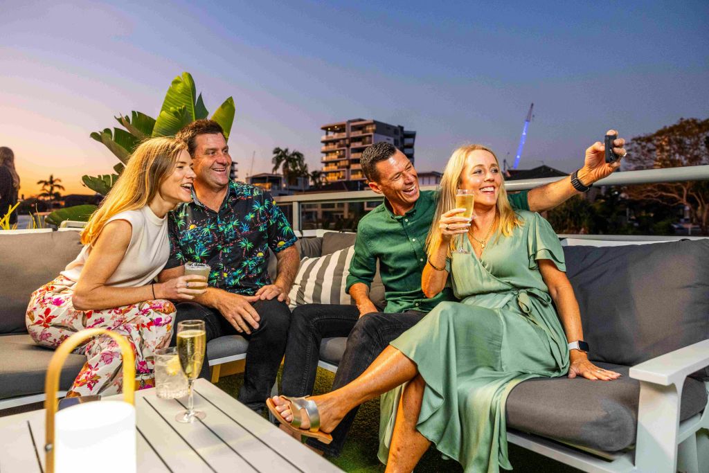 옥상 파티오에 세 사람이 앉아 있습니다. 초록색 드레스를 입은 여성이 옆에서 한 남성과 셀카를 찍고 있습니다. 도시를 배경으로 한 건물들.