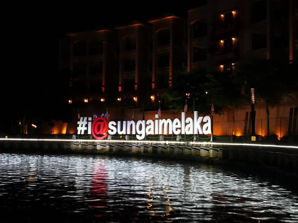 커다란 조명 간판 "# i @ sungaimelaka" 는 밤에는 바닷가에 세워져 있으며, 건물을 배경으로 조명이 켜져 있습니다.