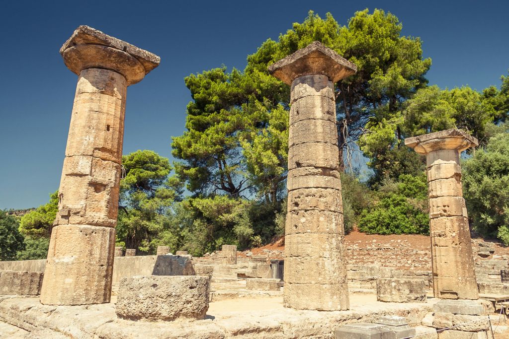 고대 석조 기둥은 맑고 푸른 하늘 아래 푸른 나무로 둘러싸인 유적지 가운데 서 있습니다.