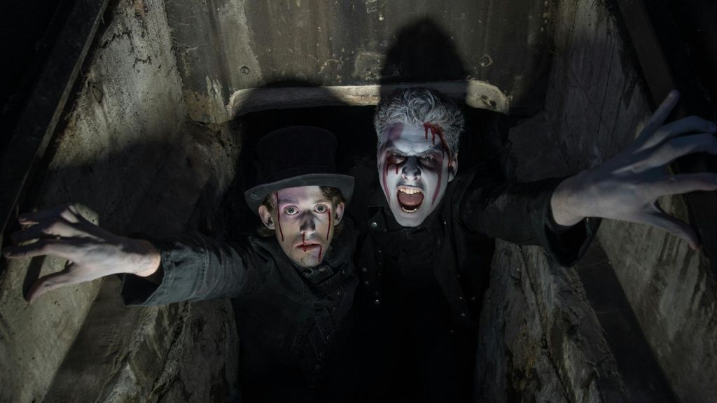 드라마틱한 메이크업과 가짜 피를 가진 두 사람이 어둡고 밀폐된 공간에서 위를 바라보고 있습니다. 한 사람은 모자를 쓰고 있습니다.