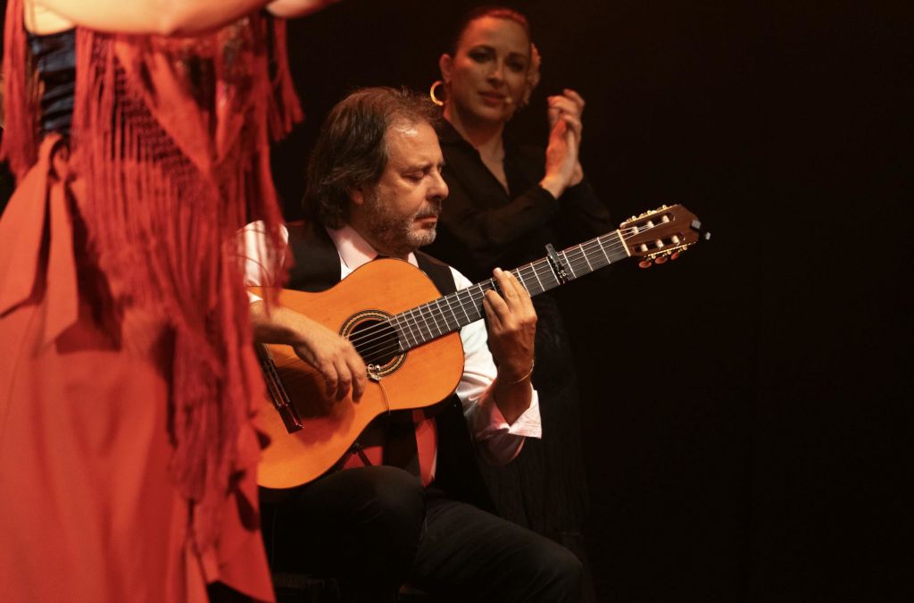 무대에서 클래식 기타를 연주하는 남성이 따뜻한 무대 조명 아래에서 여성을 배경으로 박수를 치고 있습니다.