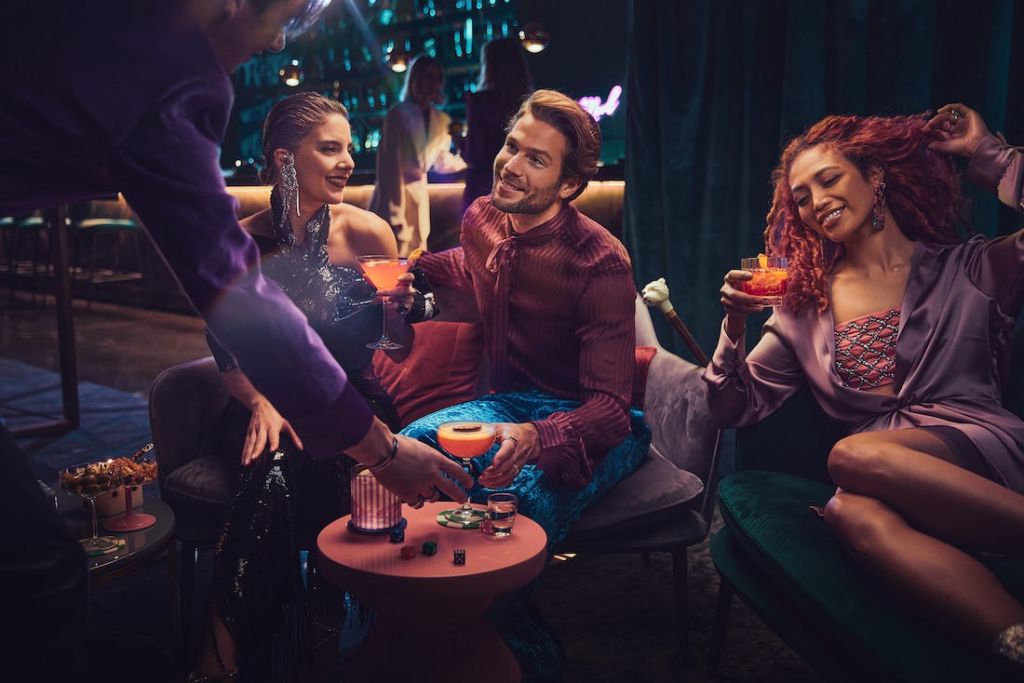 조명이 어두운 바에서 안락의자에 앉아 바텐더와 바를 배경으로 칵테일을 즐기는 세 사람.