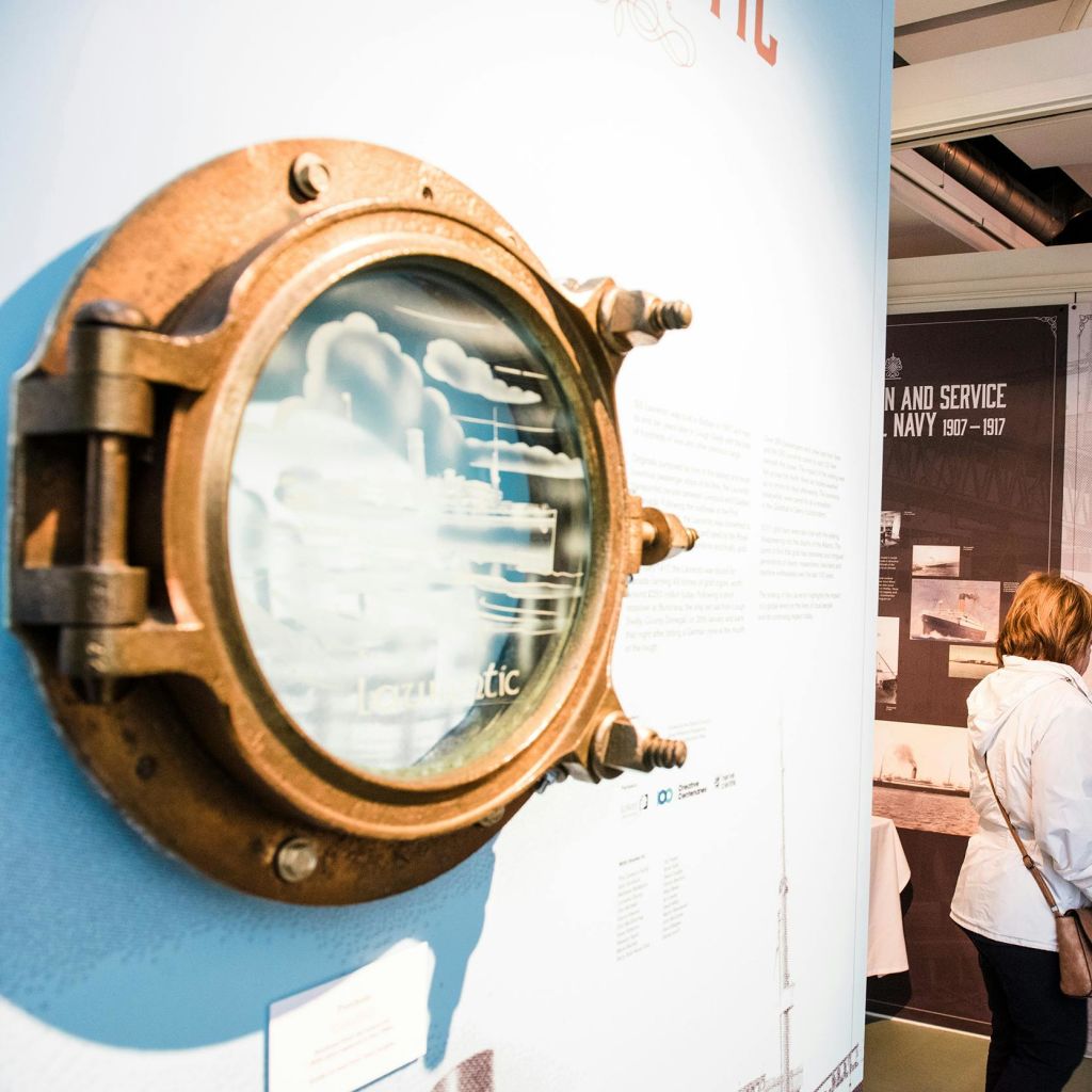 청동 선박 포트홀이 박물관 벽에 텍스트 설명과 함께 전시되어 있습니다. 흰 외투를 입은 사람이 근처의 전시물을 살펴보고 있습니다.